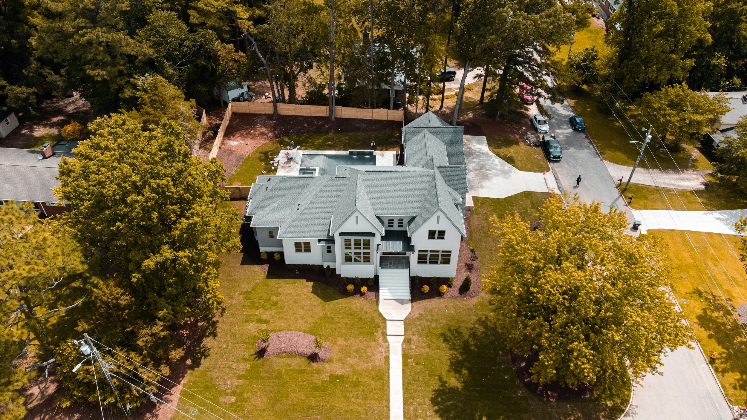 Aerial view of a white house with a gray roof, a walkway leading to the front door, surrounded by trees and a fenced backyard, with some cars parked on the street.