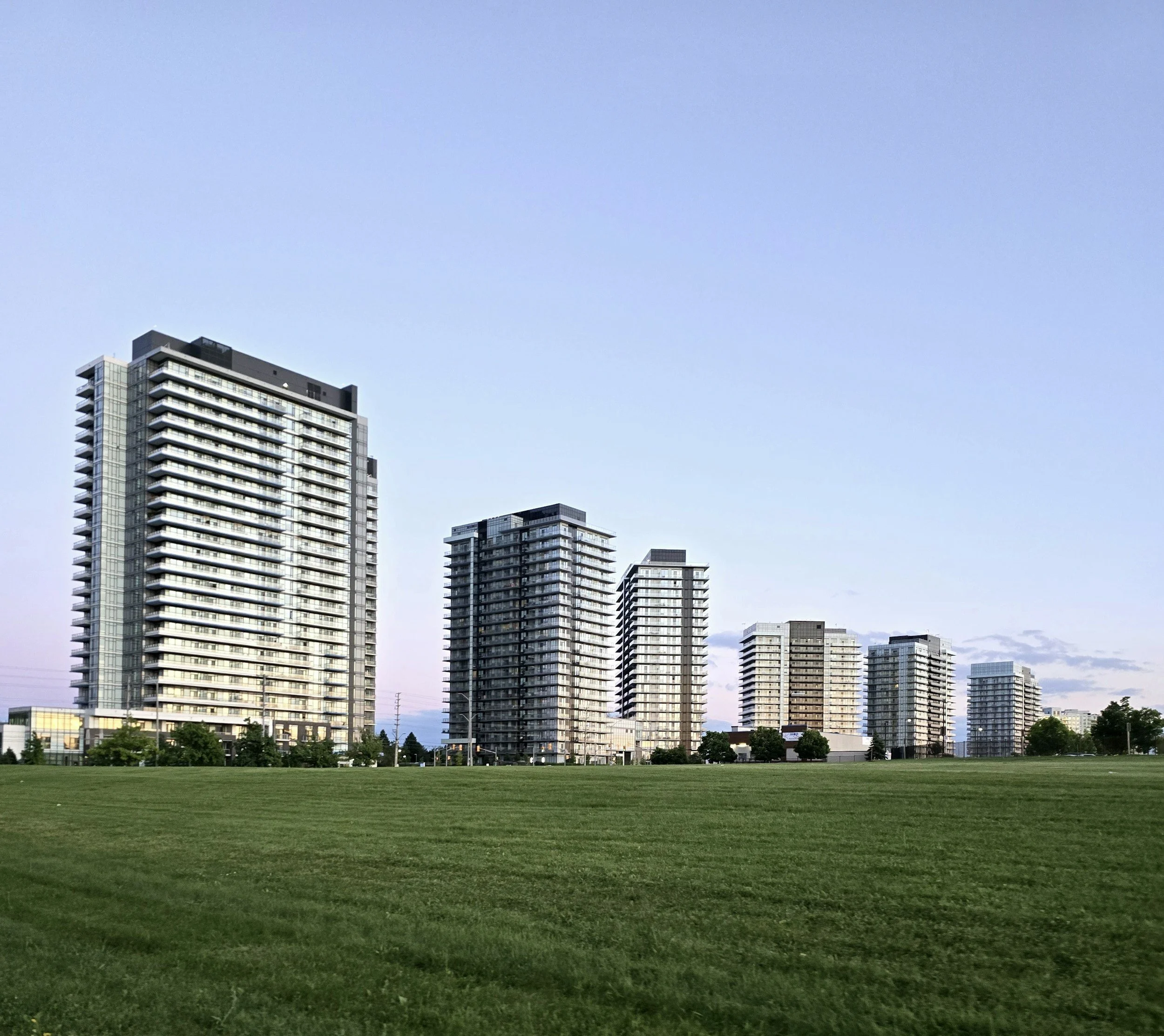 A panoramic view of a city skyline with several tall modern residential buildings made of glass and steel, set against a clear blue sky. In the foreground, there is a large, well-maintained grassy park.