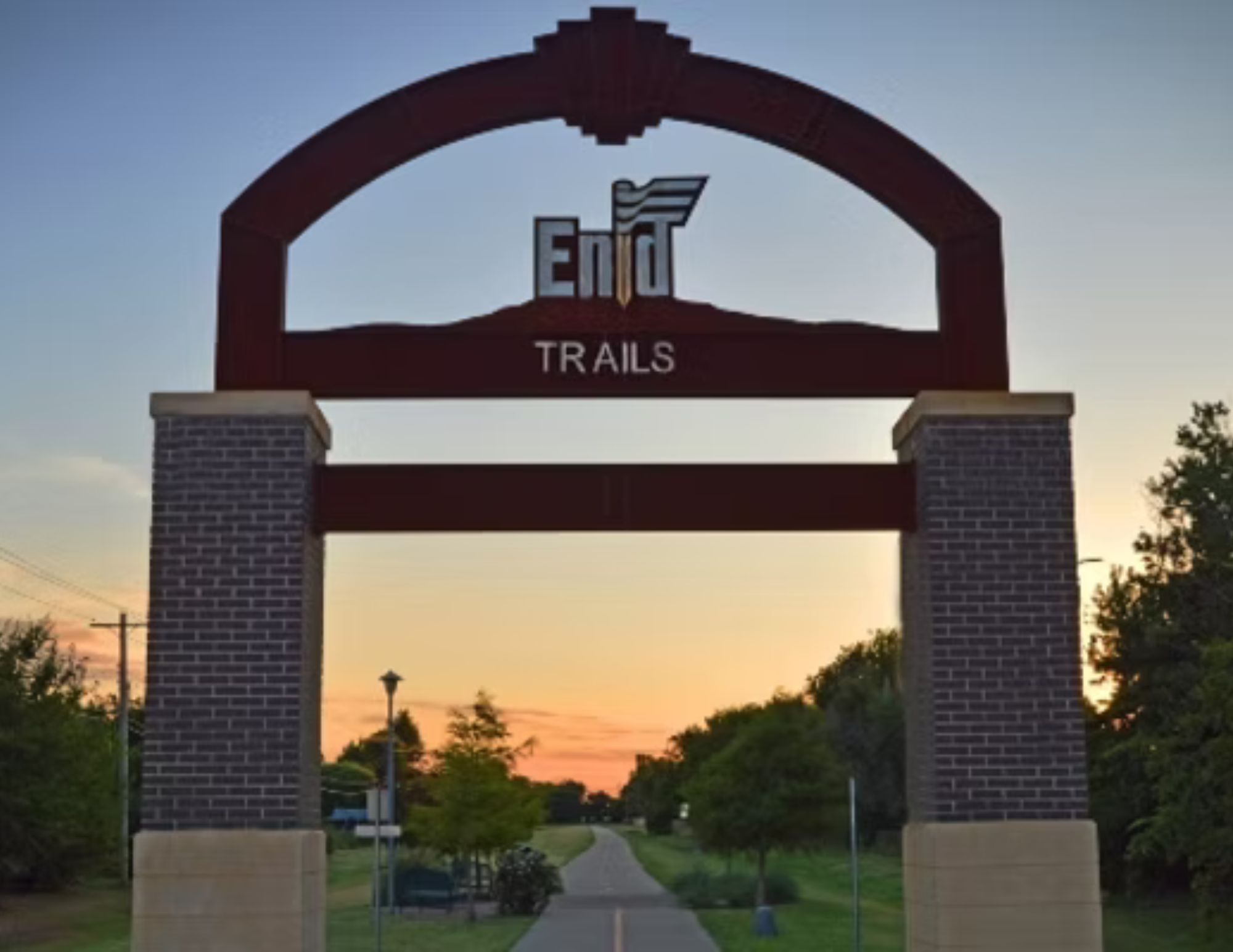 A park entrance archway with brick pillars, featuring a sign that reads 'End Trails' with a mountain design, at sunset with trees and a pathway visible in the background.