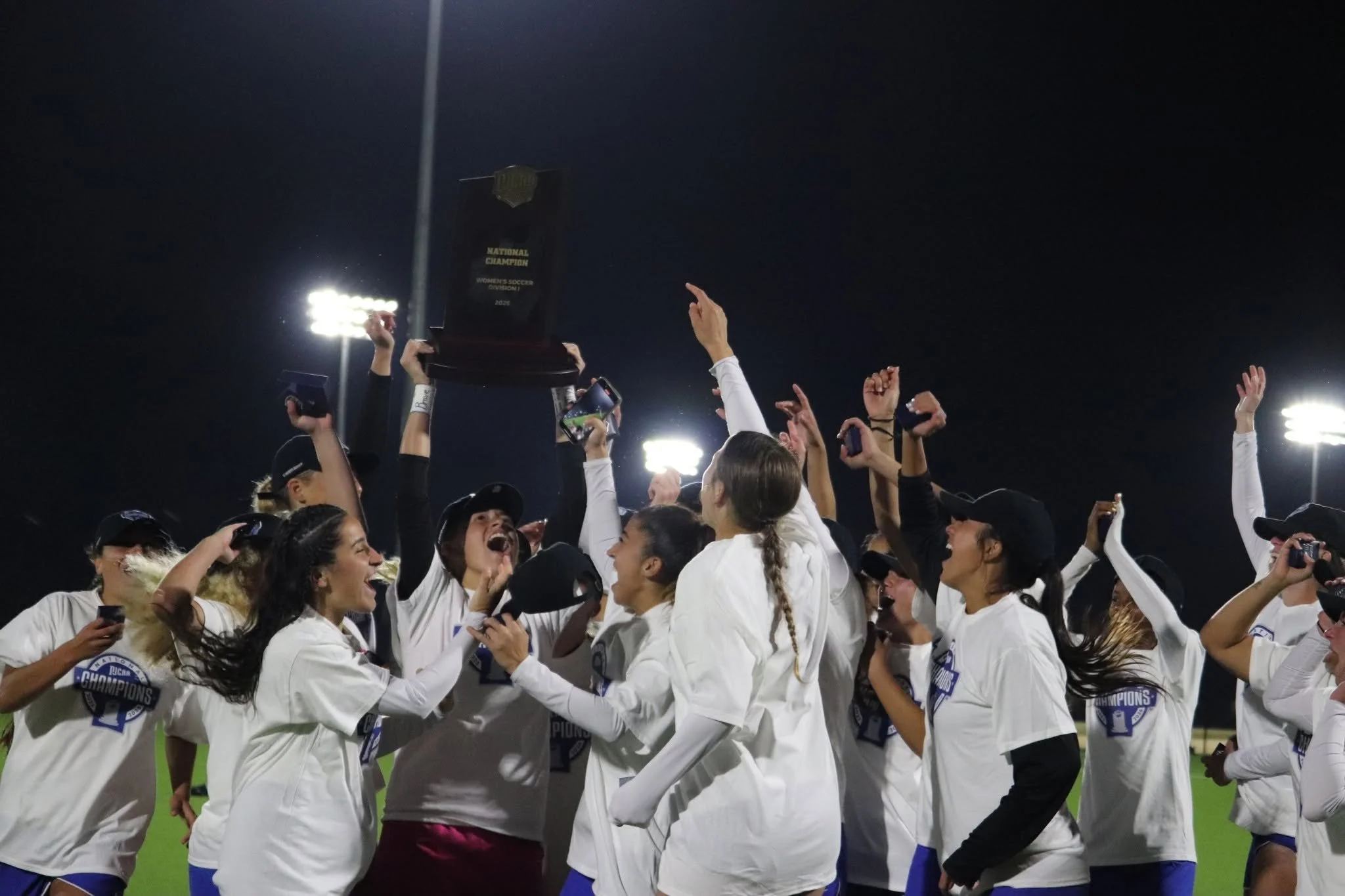 A group of female soccer players celebrating on the field at night, holding up a trophy that reads 'National Champion Women's Soccer Division I 2023', with stadium lights shining in the background.