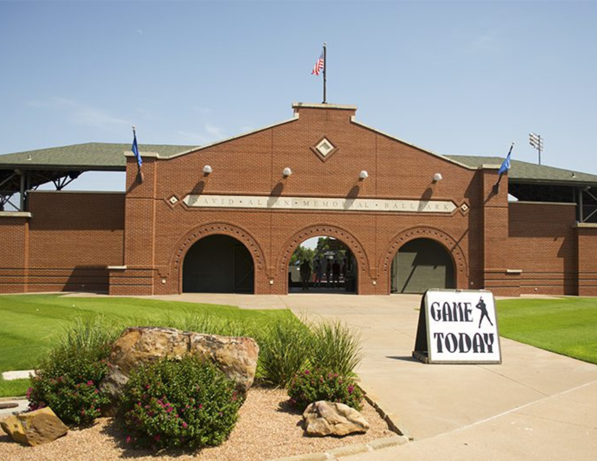 Entrance to a baseball stadium with a brick archway, a sign reading 'GAMES TODAY,' and a small flower bed in the foreground.