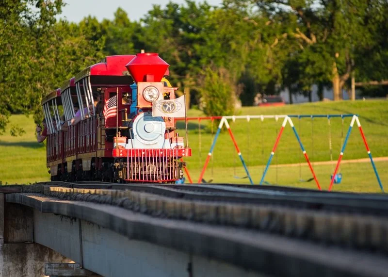 Children riding a small red train on a track in a park, with colorful swing set and green trees in the background.