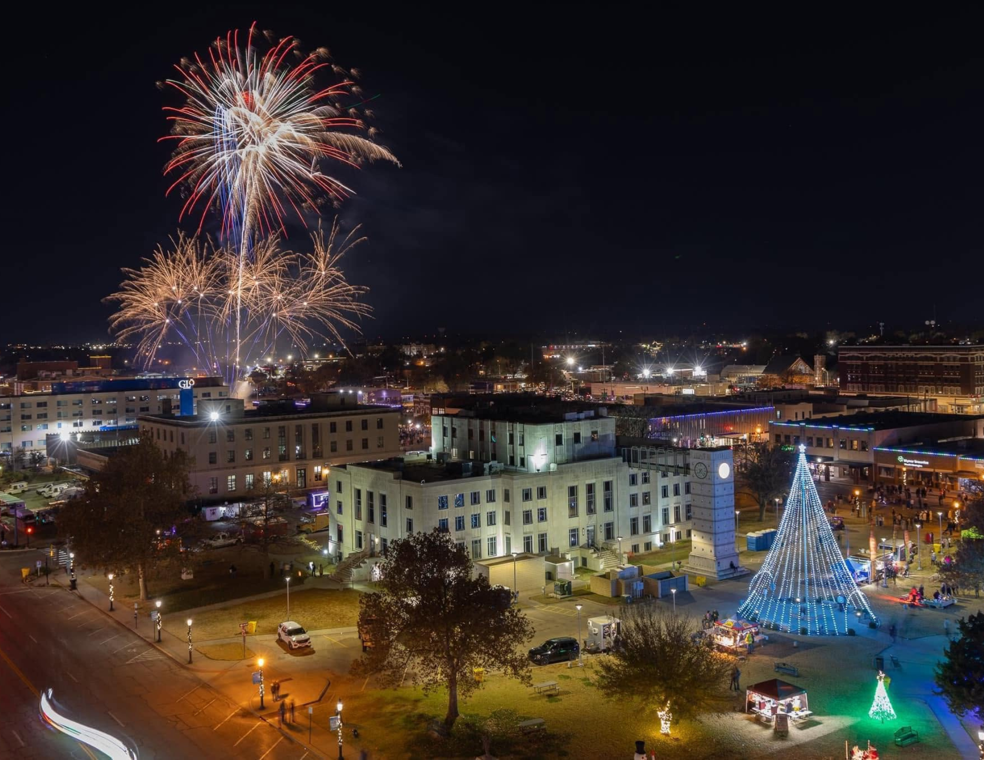 Nighttime cityscape with fireworks display and a large illuminated structure resembling a Christmas tree in the downtown area.