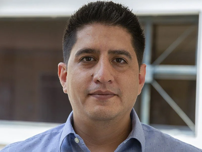 Un hombre con cabello corto y oscuro, ropa casual, fondo con estructura metálica y madera.