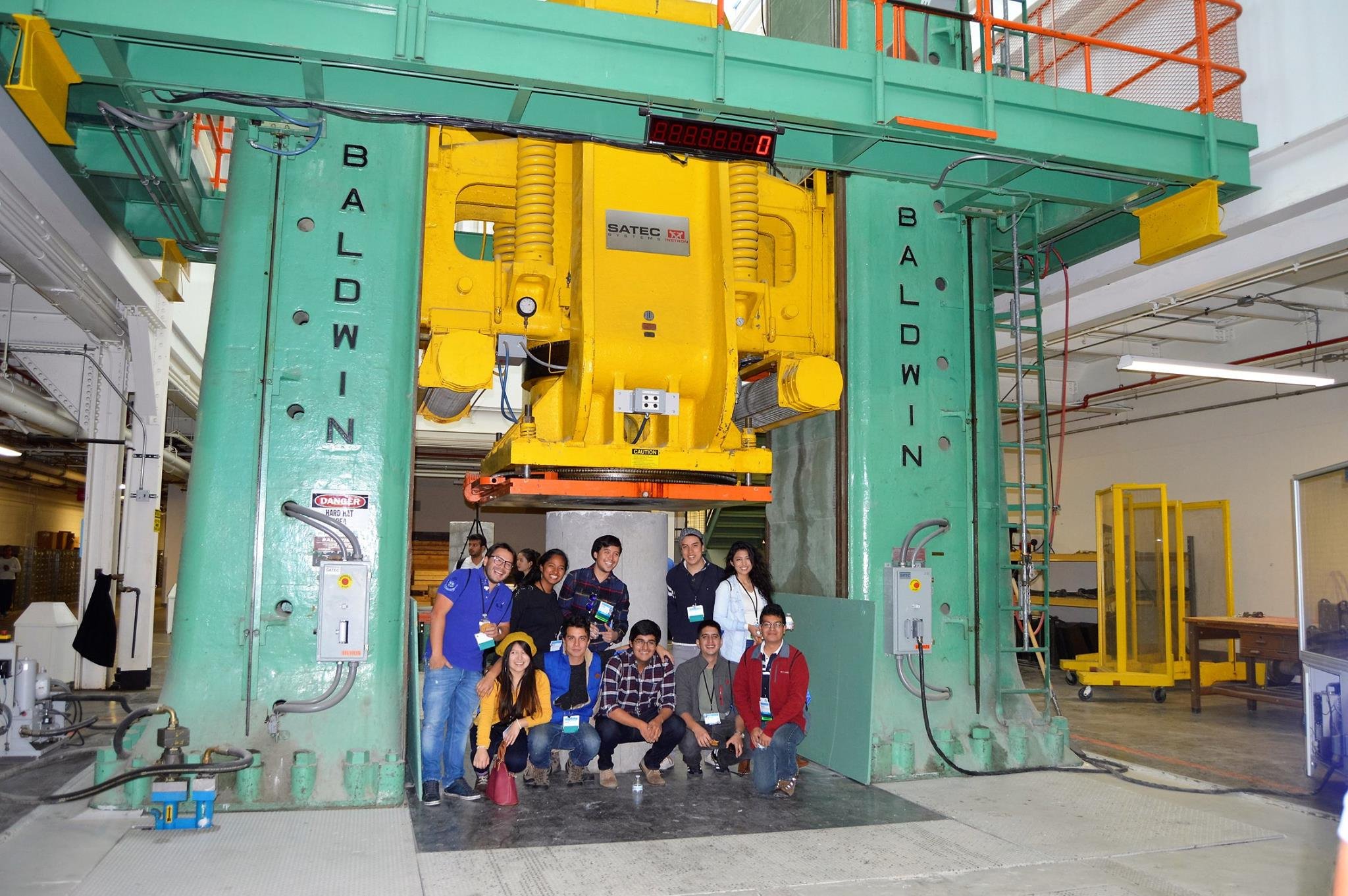 Grupo de personas posando debajo de una estructura industrial grande en una fábrica con equipo verde y amarillo.