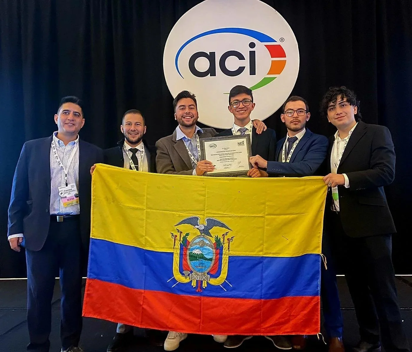Grupo de jóvenes en trajes formales con acreditaciones, sosteniendo un certificado y una bandera de Ecuador, en un evento con logotipo de ACI en el fondo.