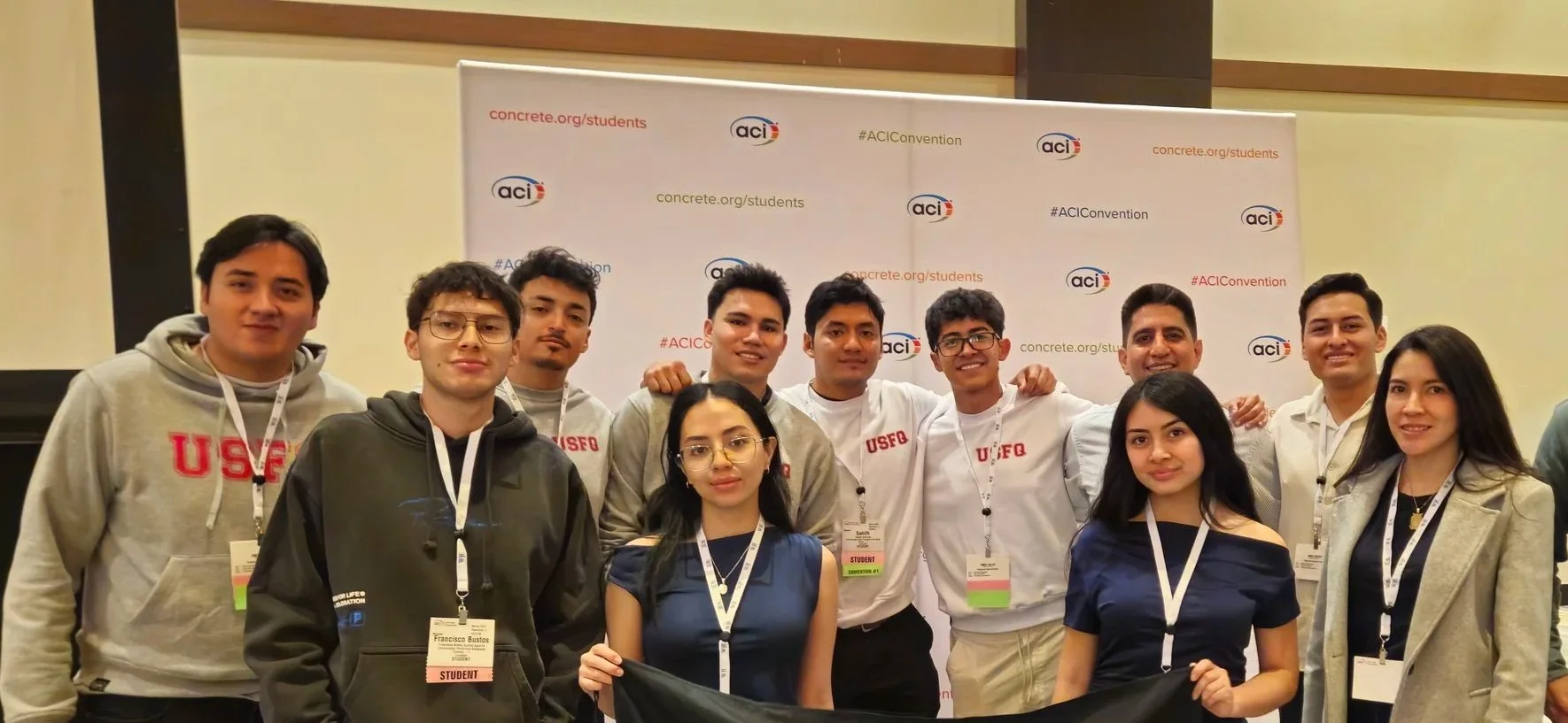 Grupo de jóvenes en un evento de la organización ACI, con un fondo que muestra logotipos y enlaces de la organización, algunos de los jóvenes llevan gafas y la mayoría usa credenciales colgadas al cuello.