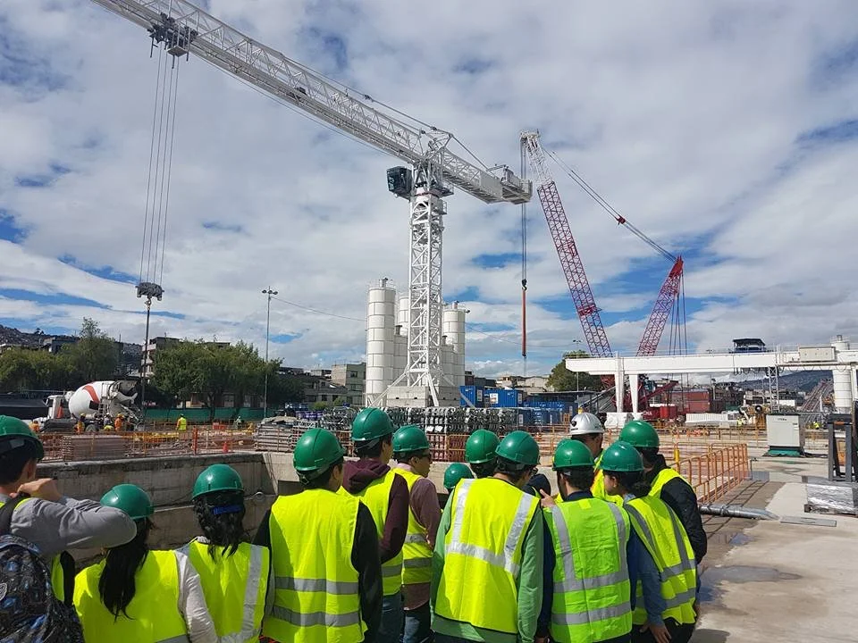 Grupo de personas con cascos de seguridad y chalecos amarillos en un sitio de construcción con grúas y maquinaria pesada.