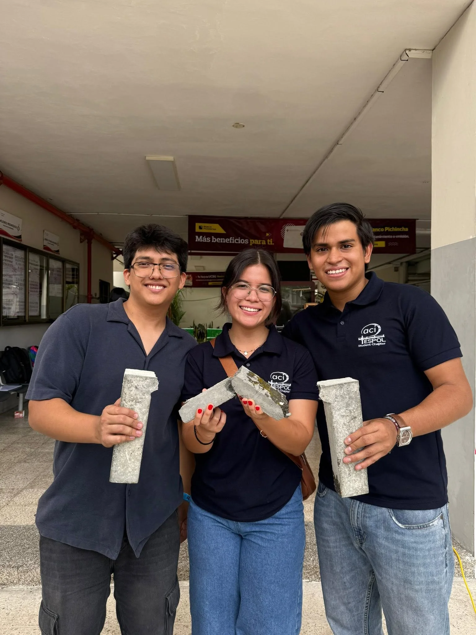Tres jóvenes estudiantes de pie en un campus, sosteniendo bloques de cemento y sonriendo, usando camisetas con el logo del club estudiantil TESPOL.