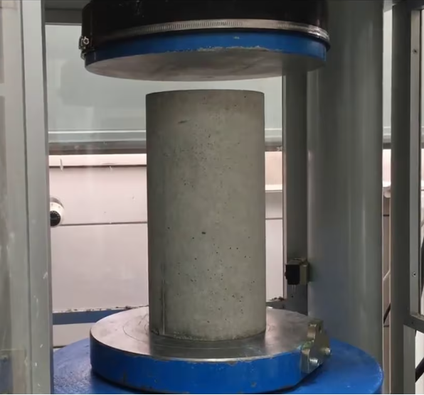 Foto de una máquina de compresión que aplica presión a un cilindro de concreto en un entorno industrial.