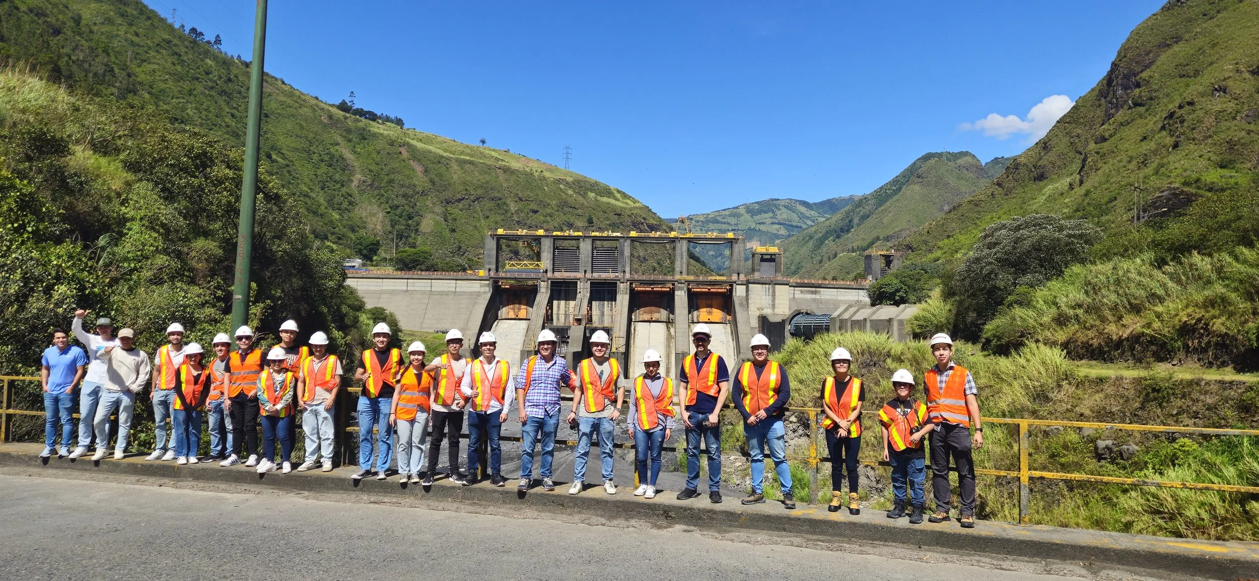 Grupo de personas con cascos y chalecos de seguridad posando frente a una presa en un paisaje montañoso con cielo despejado.