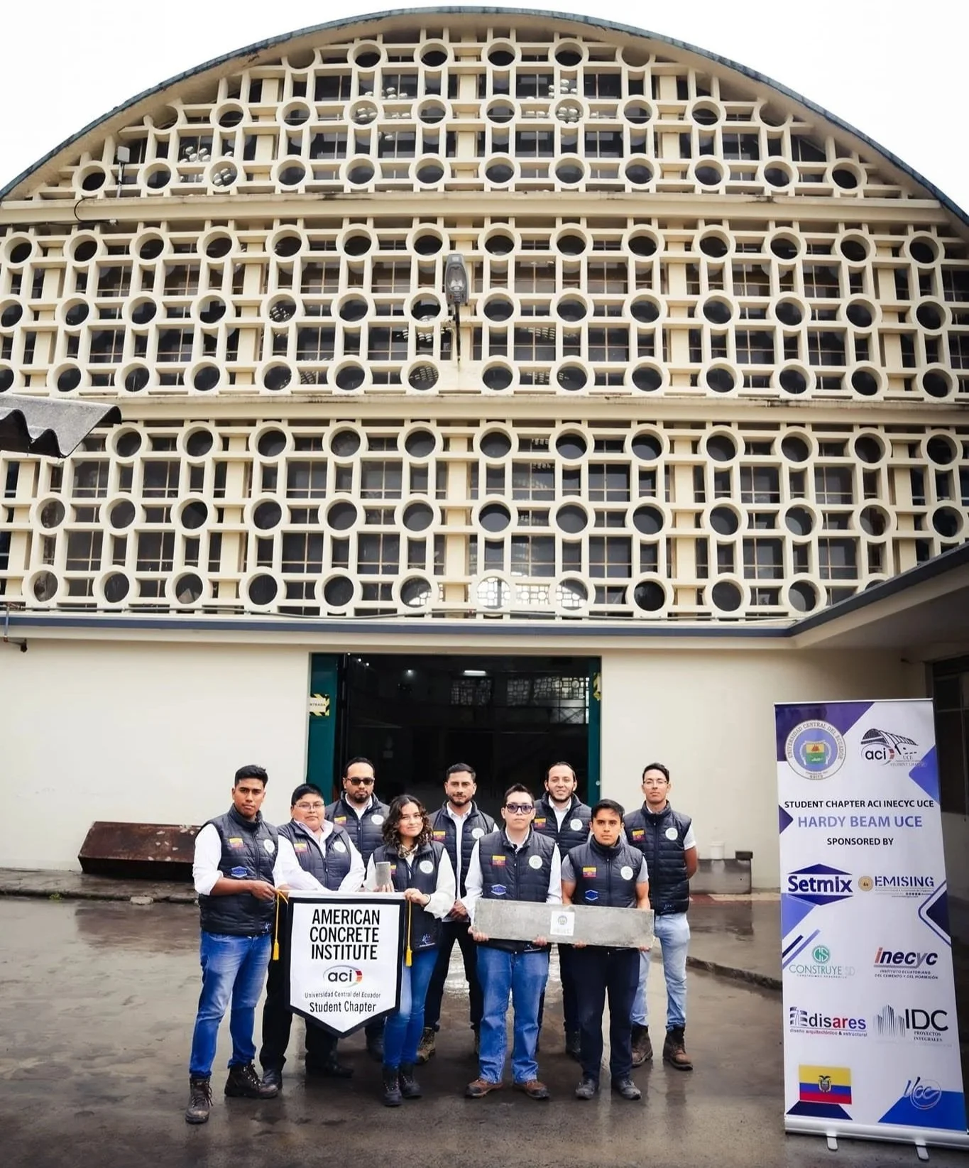 Grupo de estudiantes del Instituto Americano de Concreto en frente de un edificio con estructura de bloques de concreto. Sostienen un cartel que dice 'American Concrete Institute, Student Chapter' y una especie de molde de concreto. El grupo posa para la foto en un día nublado.