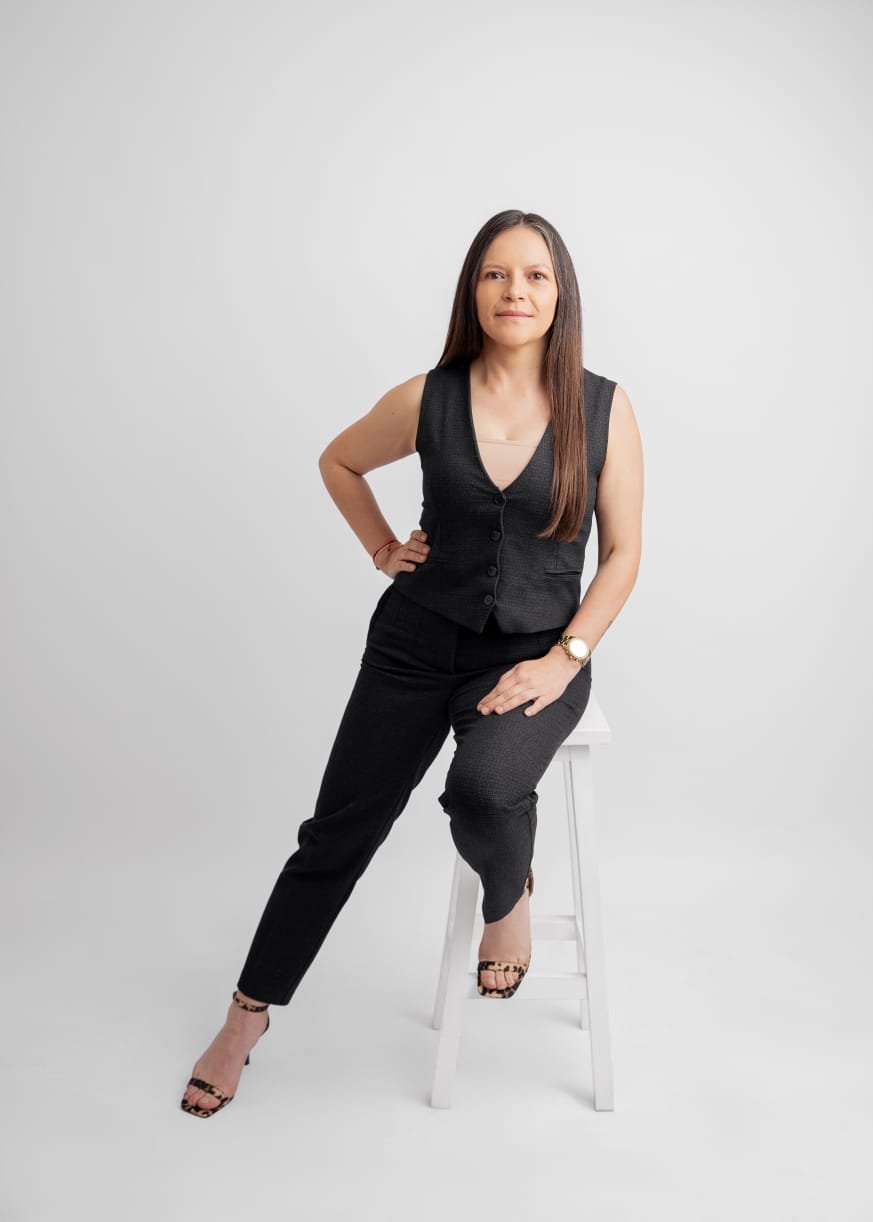 Mujer de cabello largo, vestida con ropa negra, posando en un estudio con fondo blanco, apoyada con una mano en la cadera y la otra en una silla blanca.
