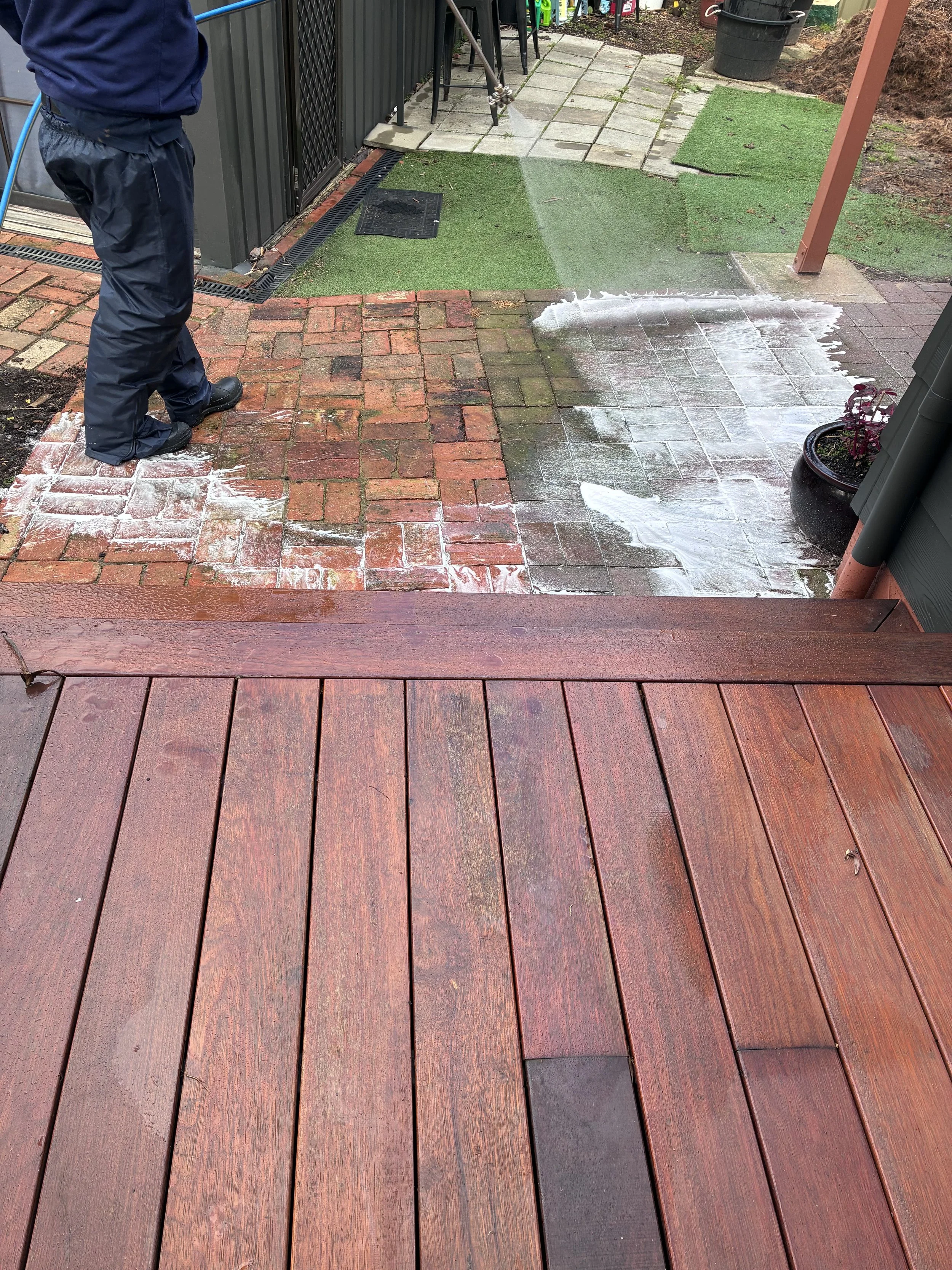 Person pressure washing a brick patio outside a building, with wooden deck in foreground, paving stones in background, and artificial grass patches.