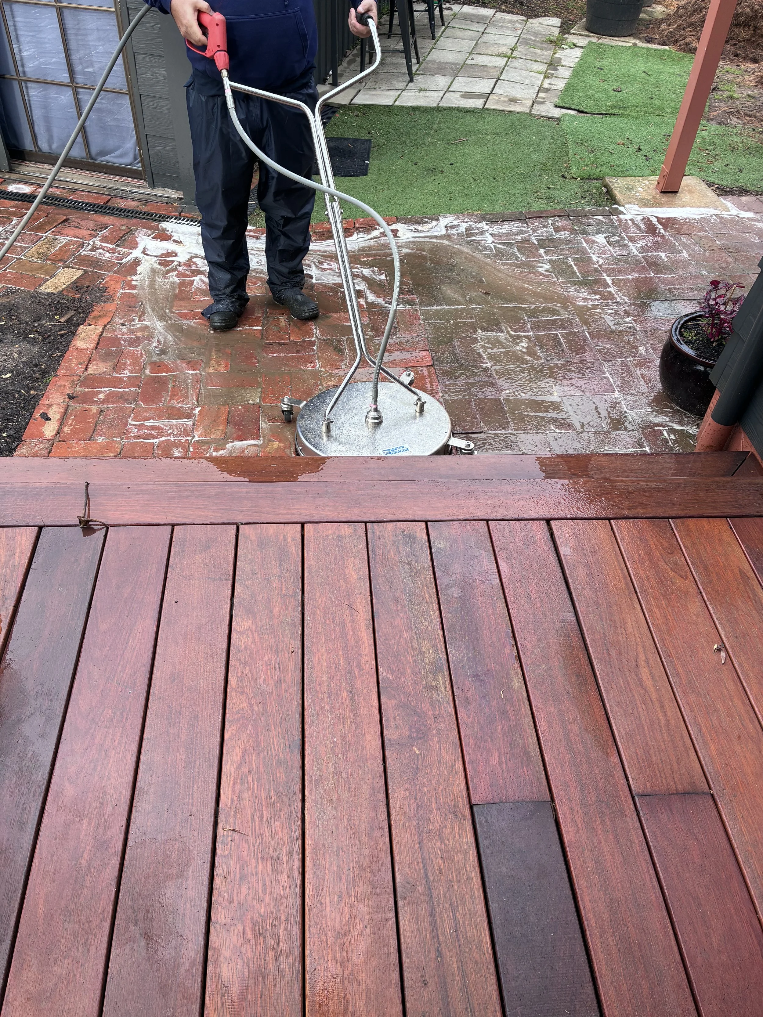 A person pressure washing a red brick patio, with water and soap visible on the surface. The person is wearing dark rain pants and shoes, and is using a red and silver pressure washer.