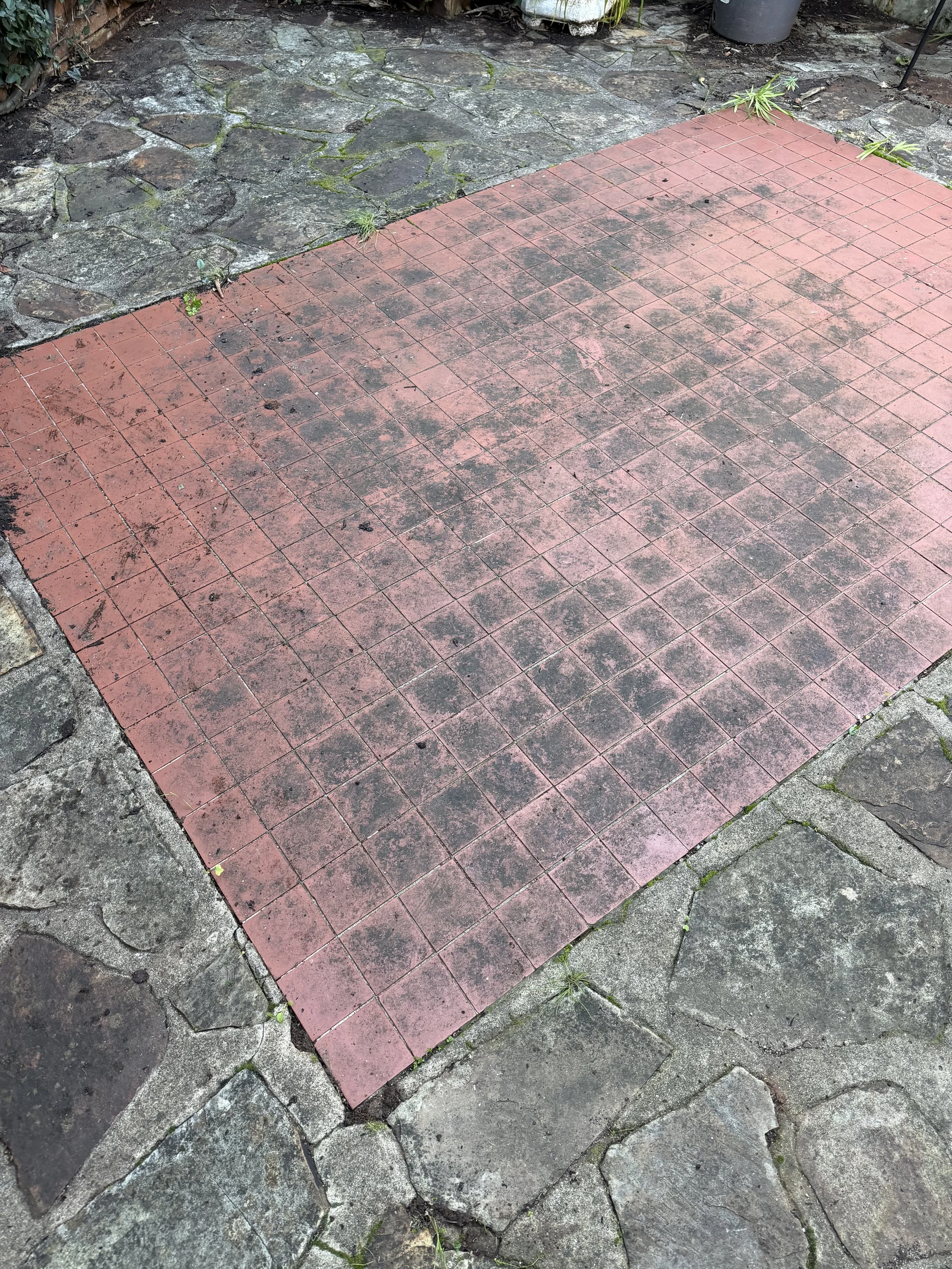 A paved outdoor area with red square tiles in the center surrounded by irregularly shaped stone slabs. in need of a cleen.