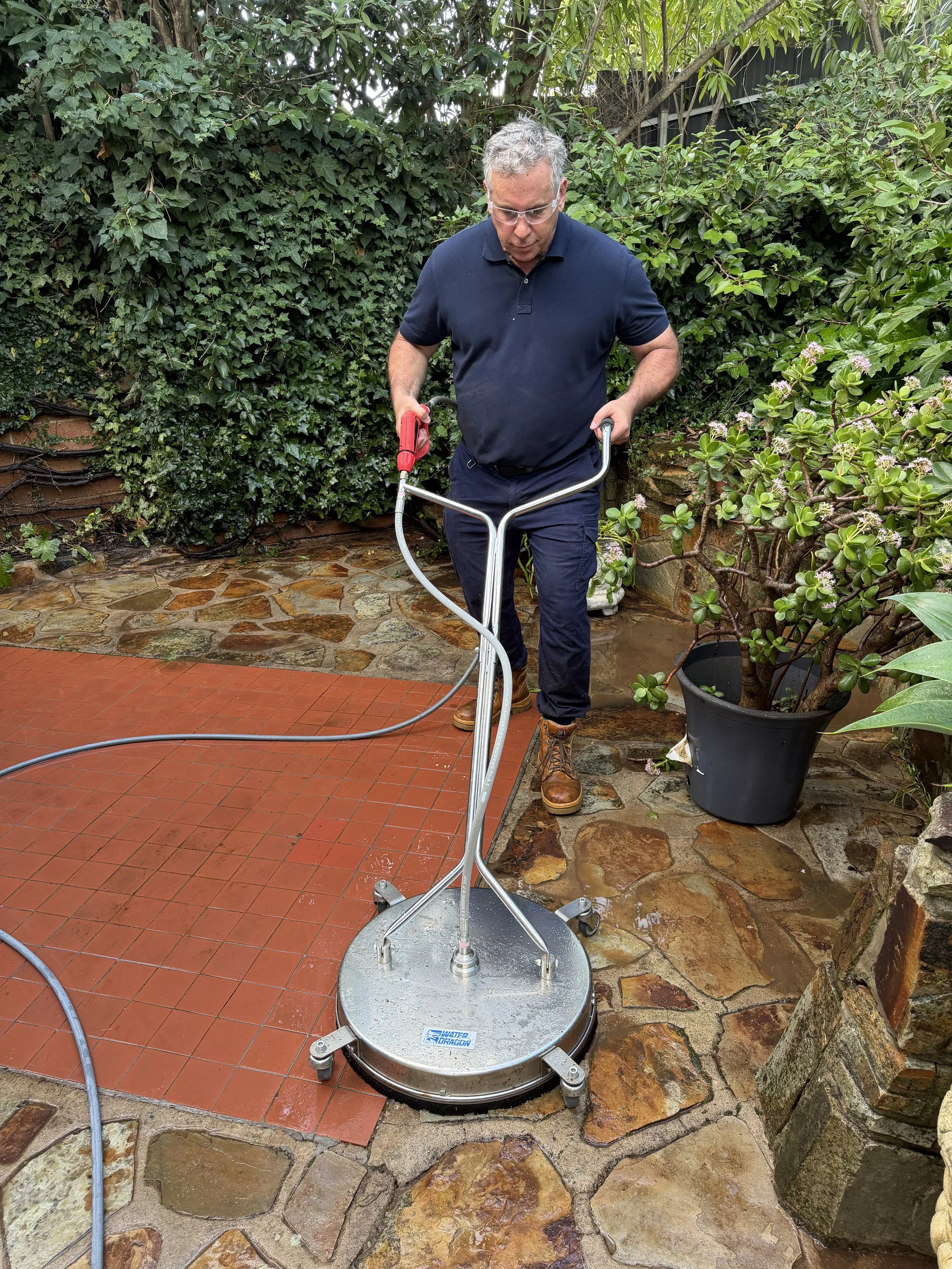Mark the owner wearing glasses and a navy blue polo shirt, operates a robotic floor scrubbing machine on a patio surrounded by green plants and bushes.