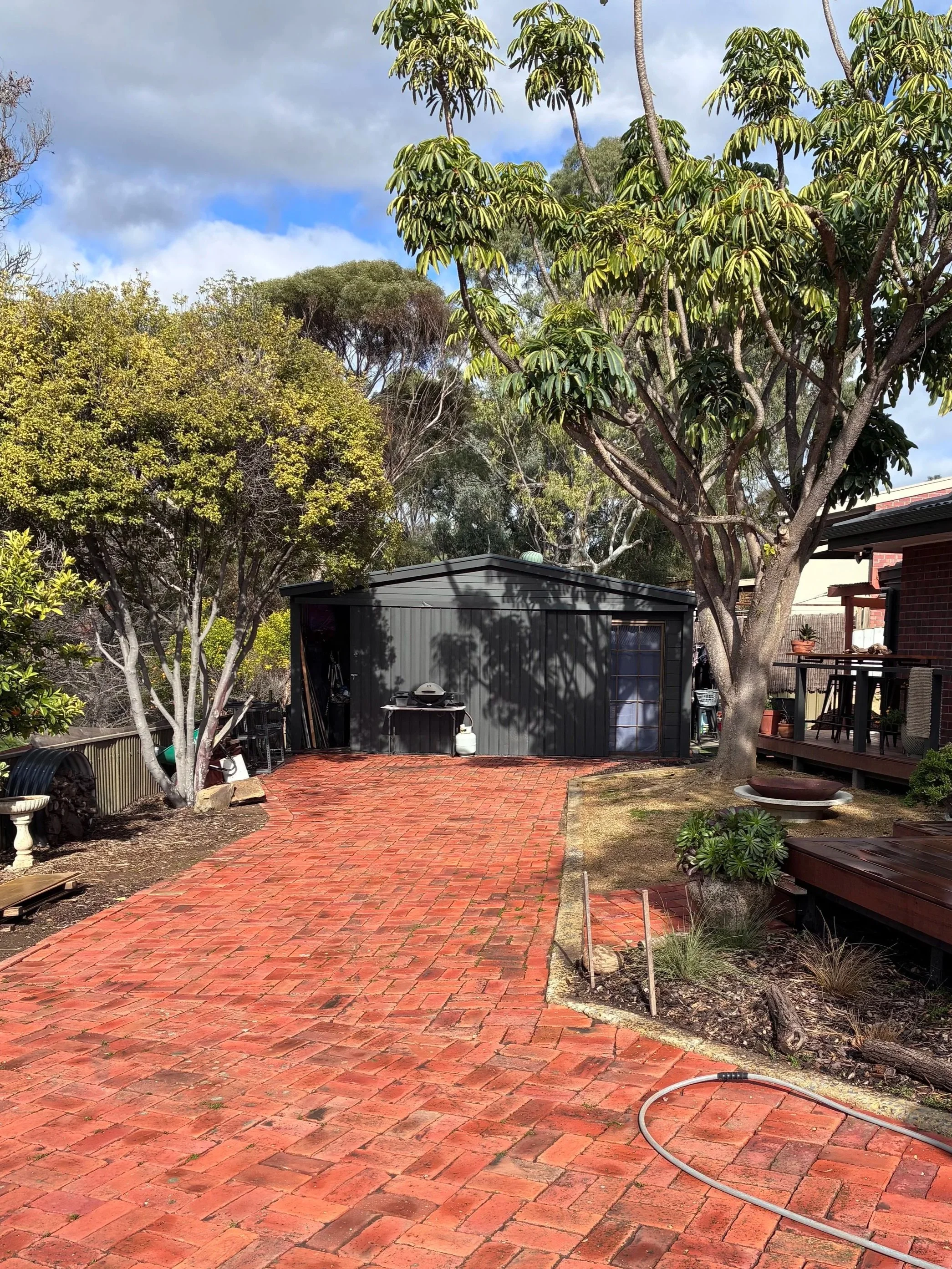A backyard patio with red brick paving, surrounded by trees and plants. There is a black outdoor shed with a barbecue grill and propane tank, and a garden hose on the ground. A wooden deck with seating is visible on the right side, and the sky is partly cloudy.