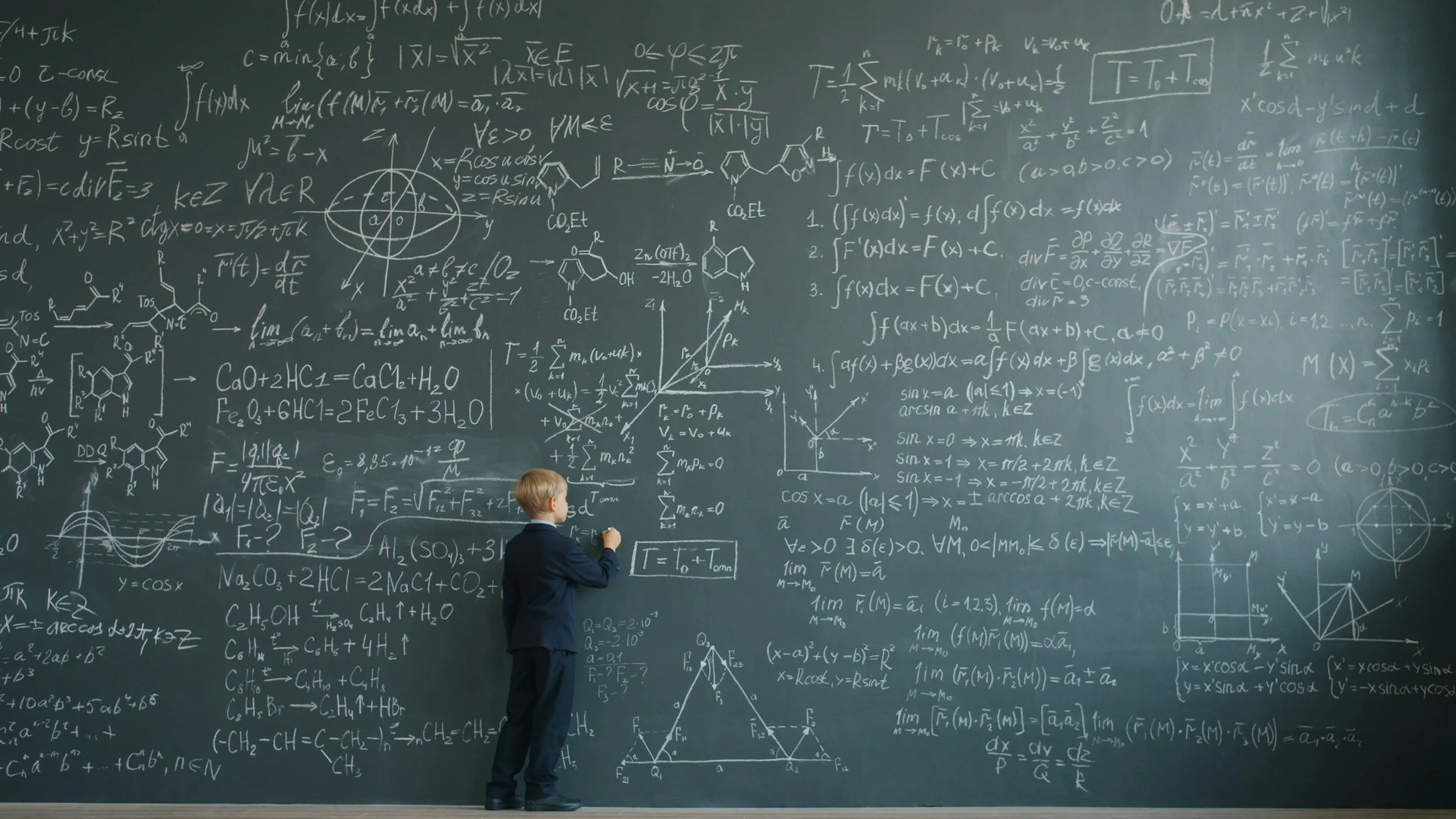 A man in a suit standing in front of a large blackboard filled with complex mathematical equations, graphs, and chemical structures.