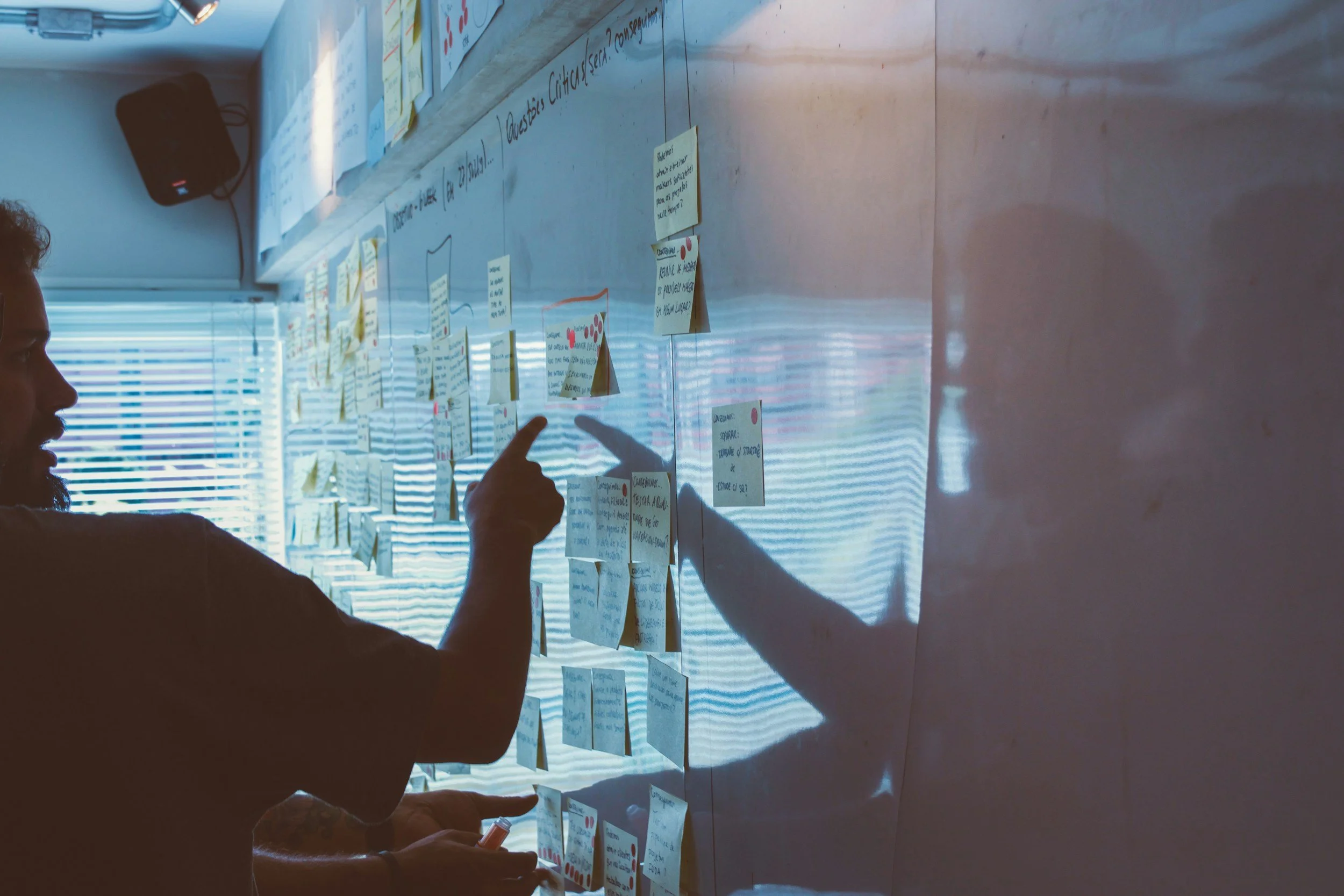 Person pointing at notes on a wall covered with sticky notes and papers, with blinds on the window and a speaker in the background.