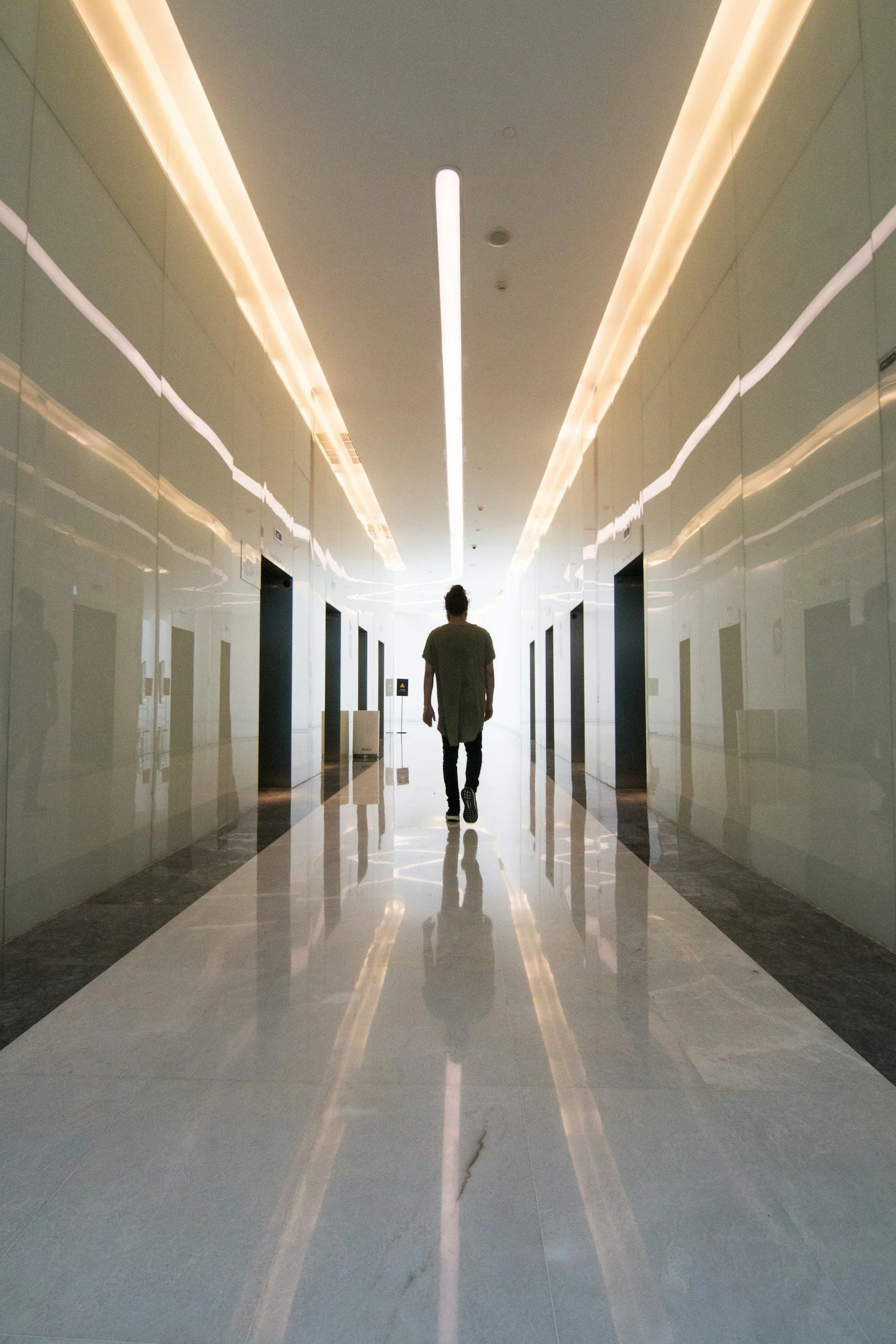 A person walking down a modern, sleek corridor with reflective walls and ceiling lighting.