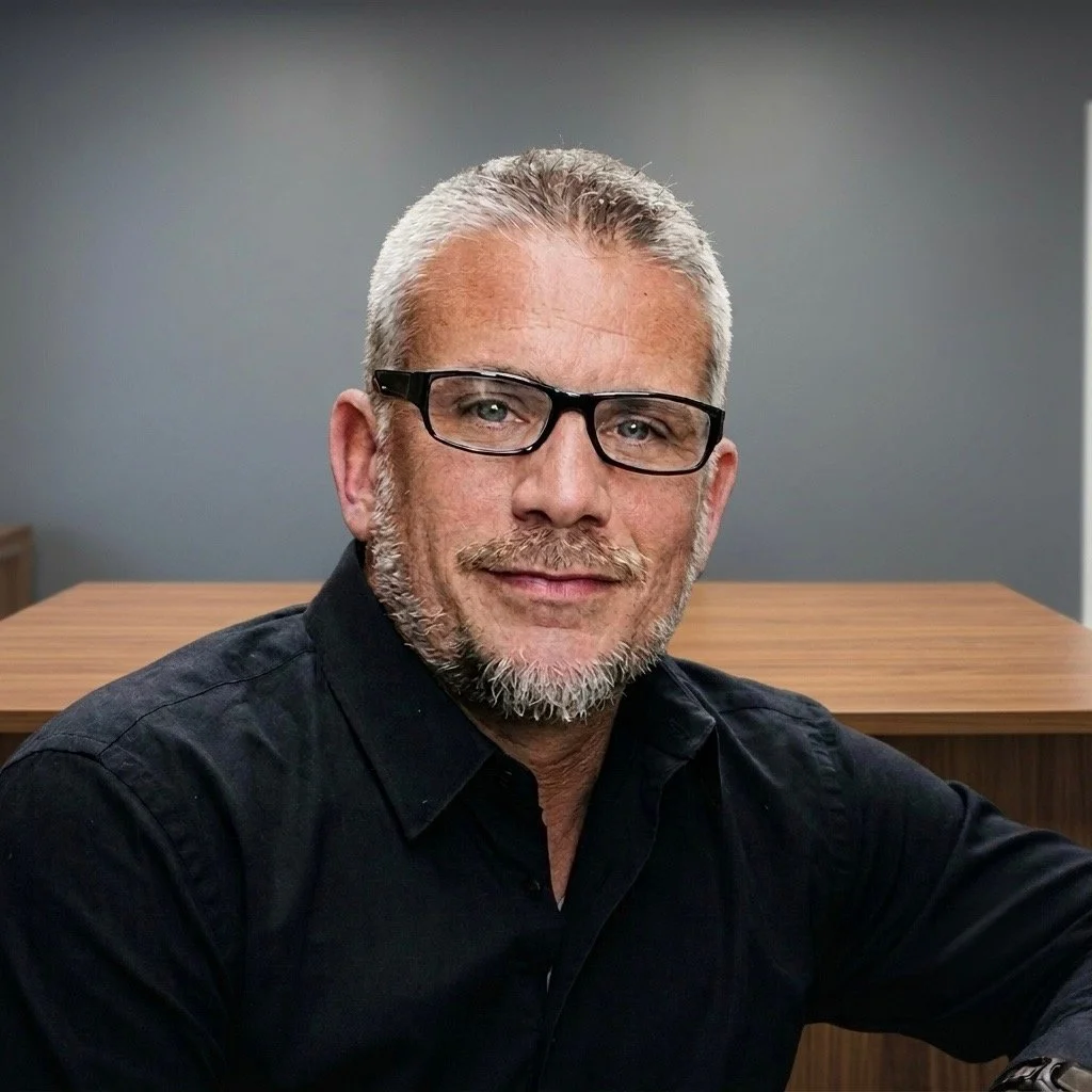 A man with gray hair, glasses, and a beard, wearing a black shirt, sitting at a wooden table with a gray wall background.