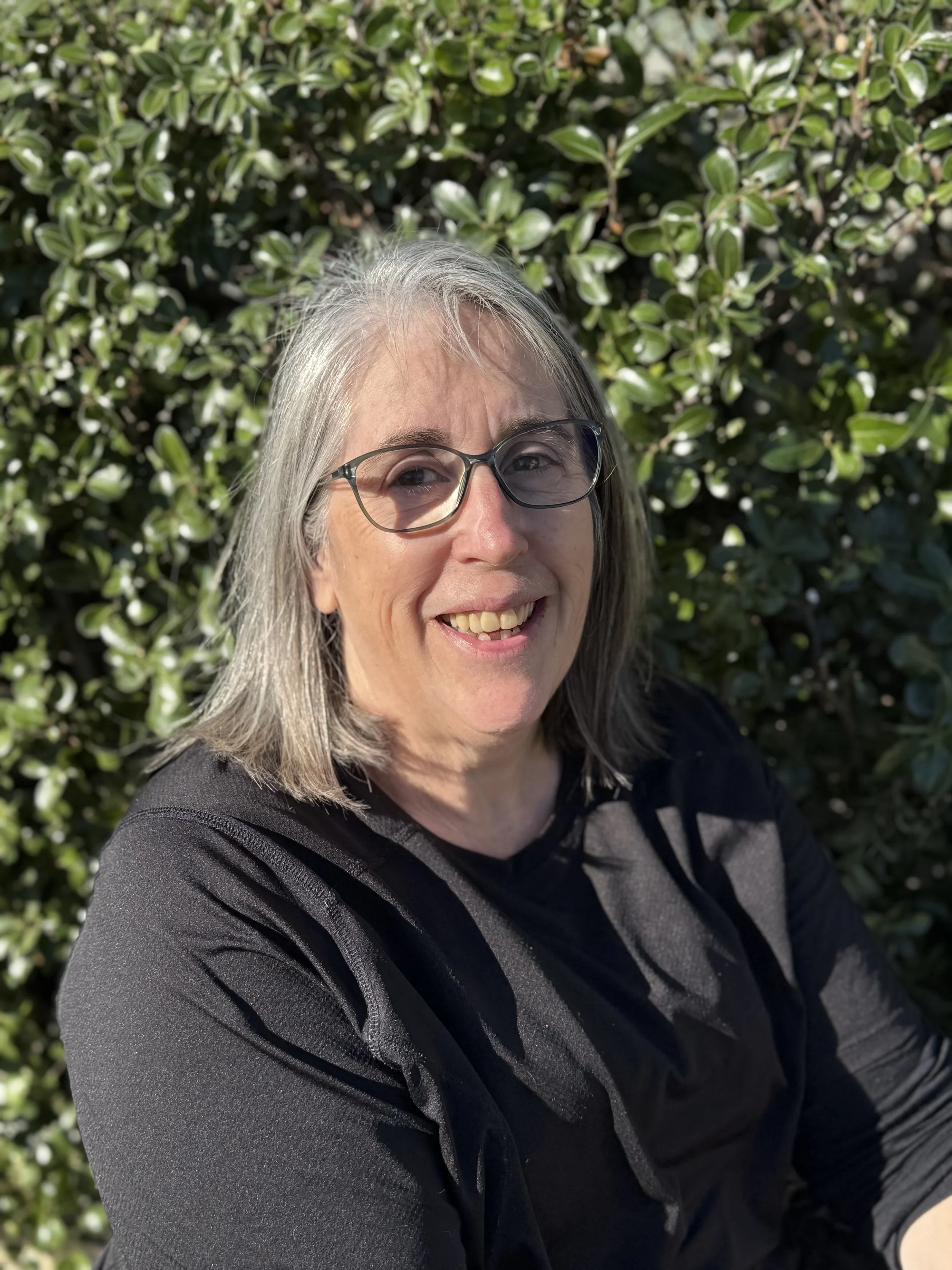 Close-up of a smiling older woman with gray hair and glasses, standing outdoors in front of a green leafy bush.