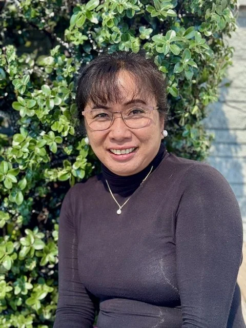 A woman wearing glasses, a necklace, and earrings, smiling while standing outdoors in front of green leafy bushes.