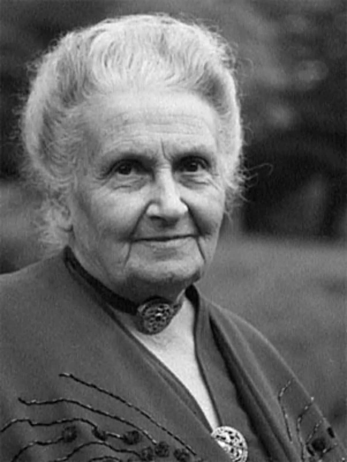 Black and white portrait of an elderly woman with short, curly hair, wearing a necklace and a patterned shawl, smiling softly outdoors.