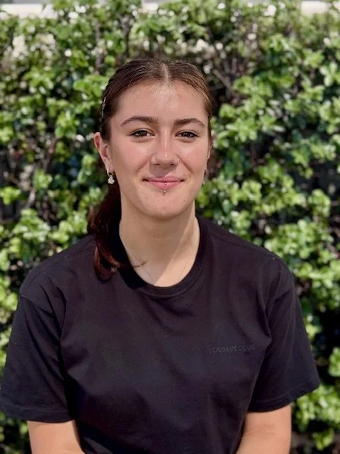 Young woman with brown hair in a braid, wearing a black t-shirt and earrings, standing in front of green bushes with white flowers.