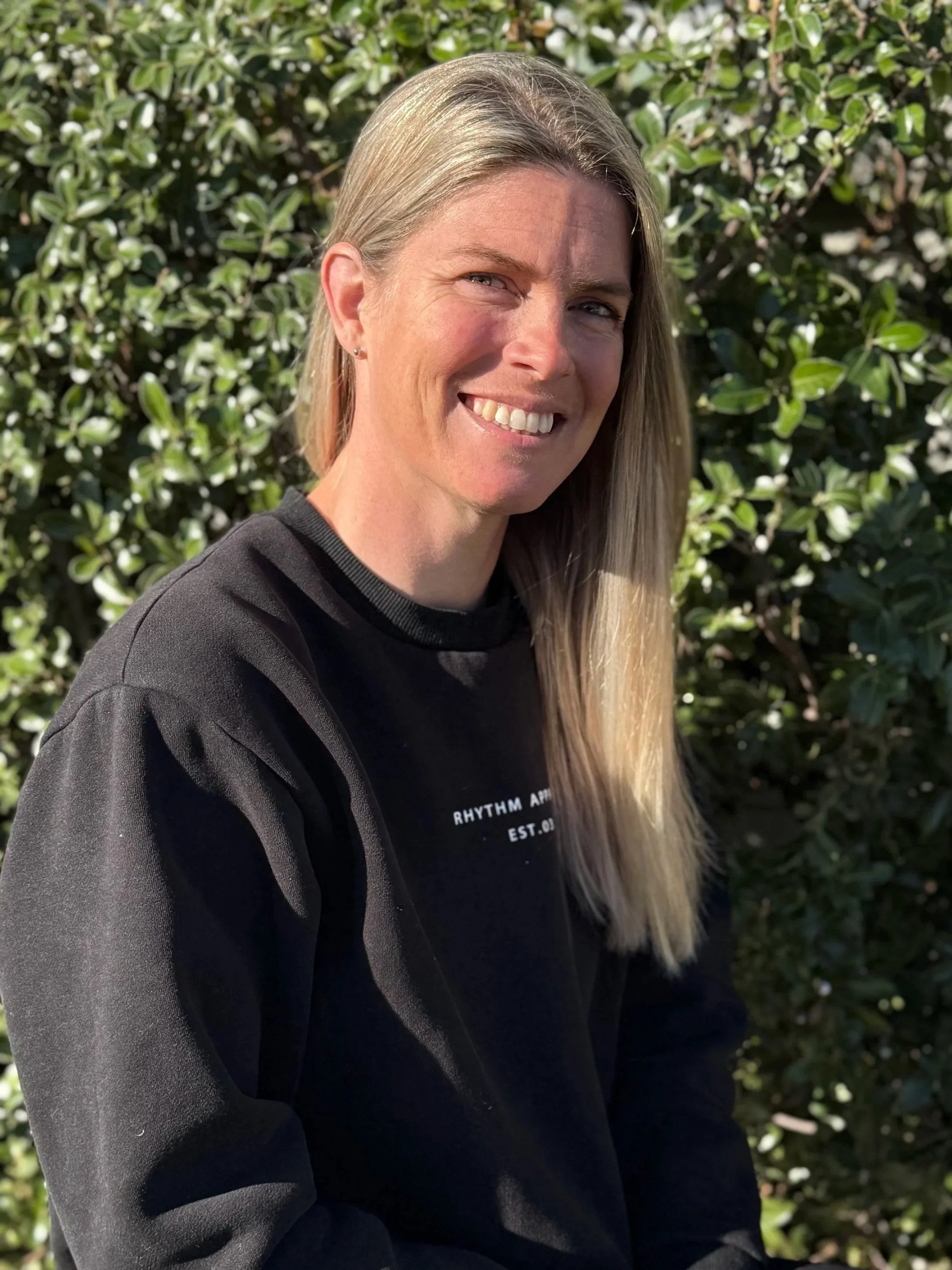 A woman with long blonde hair, smiling, standing outdoors in front of green leafy bushes, wearing a black shirt and a black jacket.