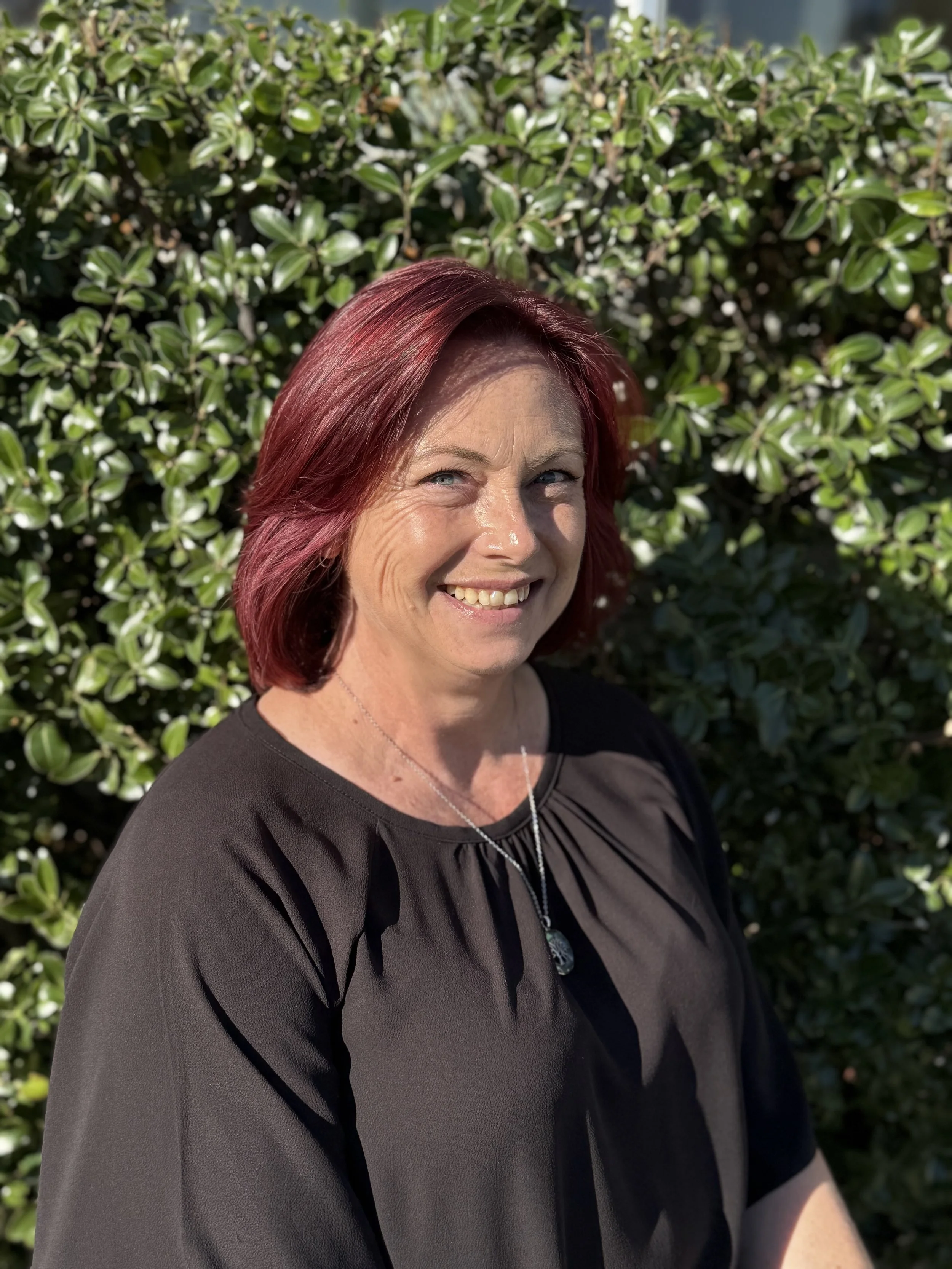 Smiling woman with red hair in front of a green leafy bush, wearing a black top and a necklace.
