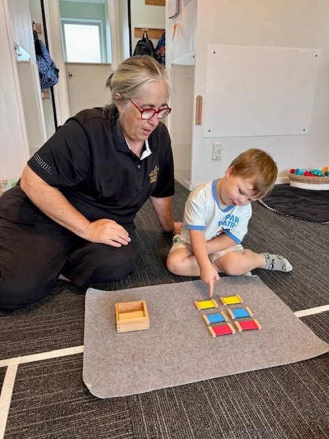 A woman and a young boy sitting on a carpeted floor, playing with colorful wooden tiles arranged on a gray mat. The woman is observing while the boy is placing a tile. The setting appears to be a classroom or playroom with a whiteboard and storage shelves in the background.