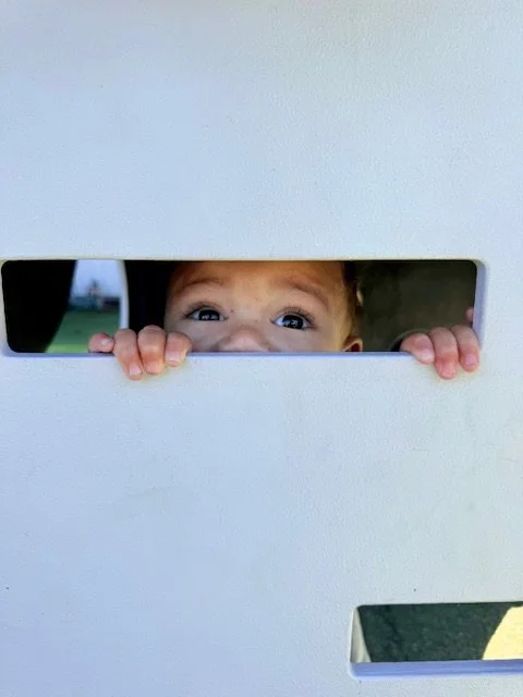 A child peeking through a rectangular hole in a white wall, with only their eyes and hands visible.