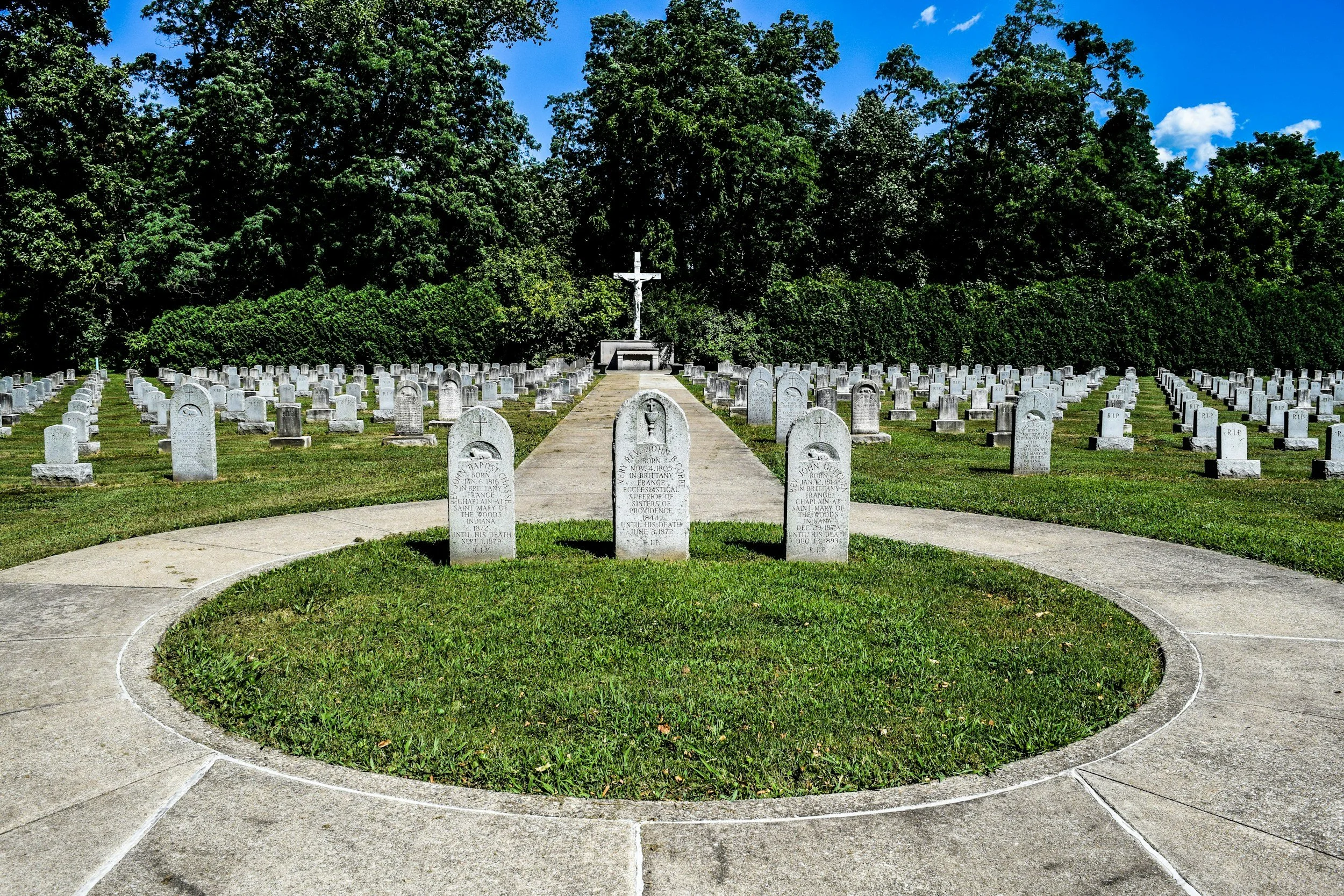 A cemetery with rows of white headstones surrounded by green grass and trees, featuring a large cross monument in the background under a blue sky with a few clouds.