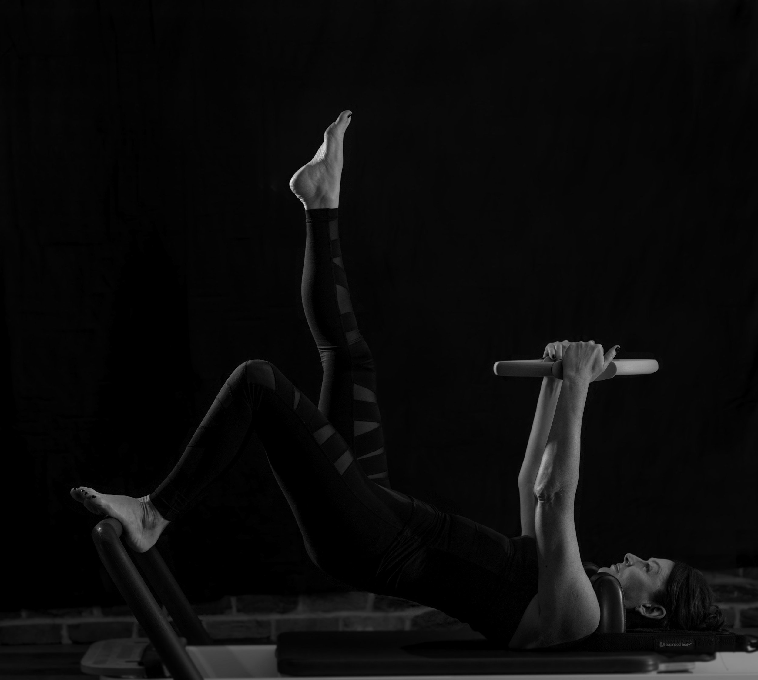 Hands with yoga or Pilates grips holding onto a horizontal bar, with legs extended upward in a stretch or pose, against a black background.