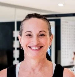 Close-up of a smiling woman with dark hair tied back, in a fitness or gym environment.