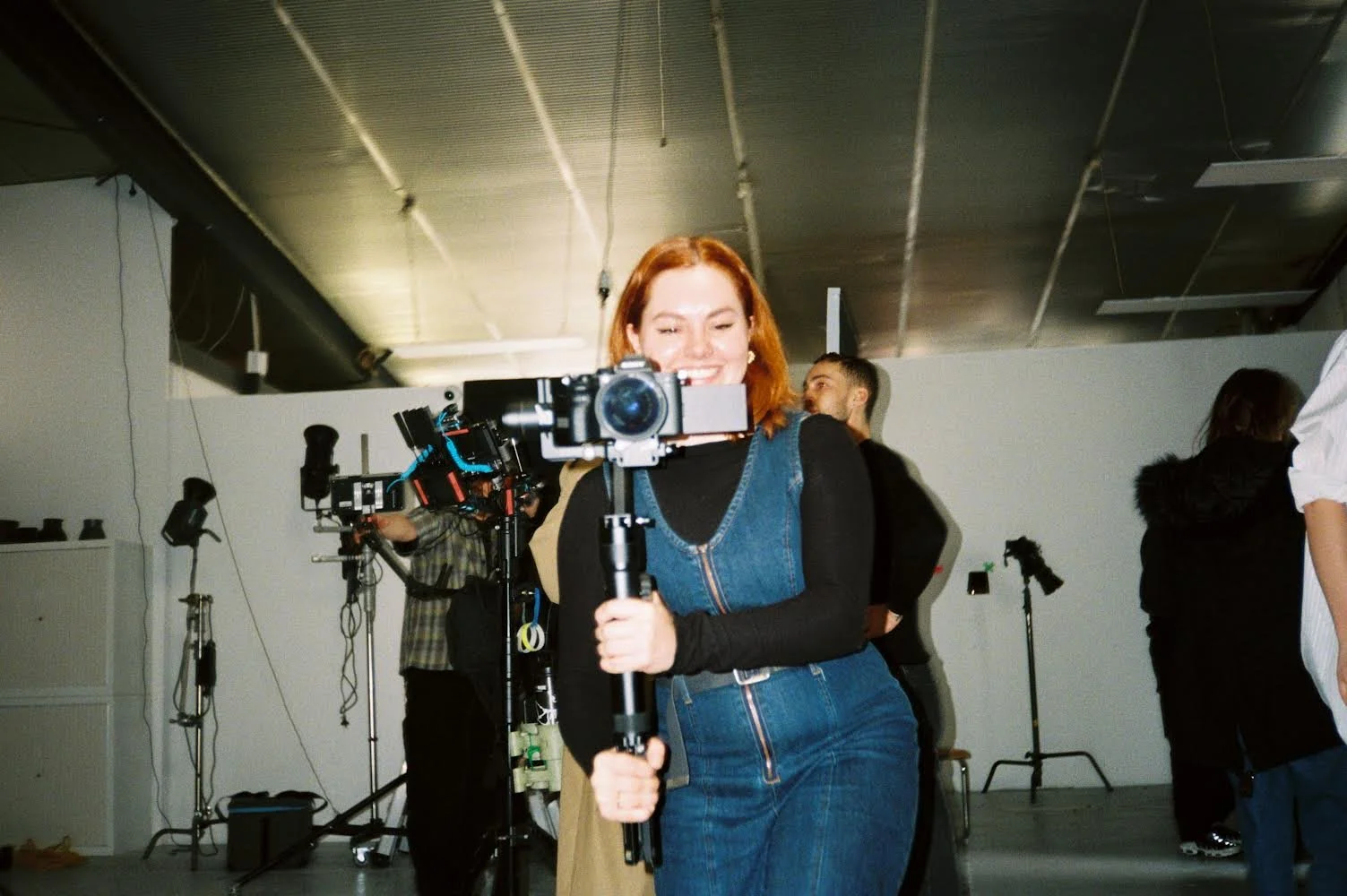 A woman with red hair smiling while holding a camera on a stabilizer in a photography studio with other people and equipment in the background.