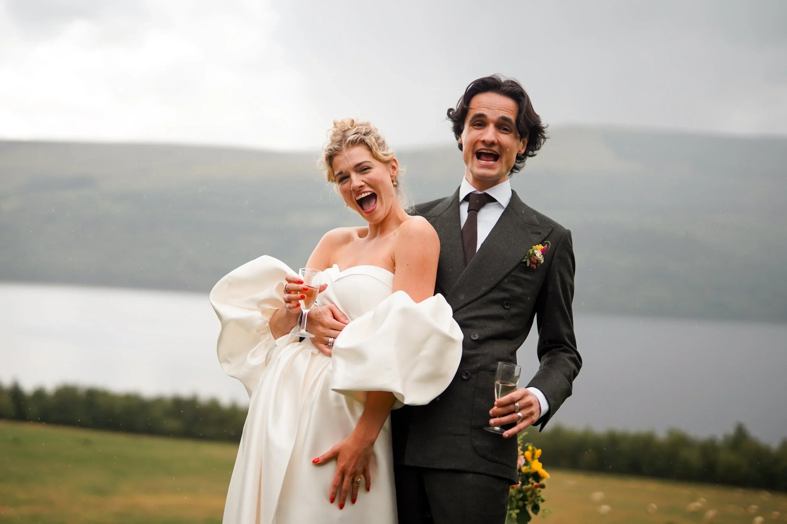 A newlywed couple smiling and celebrating outdoors on their wedding day, with the woman wearing a white wedding dress and the man in a dark suit holding a glass of champagne. The background features an open field and cloudy sky.