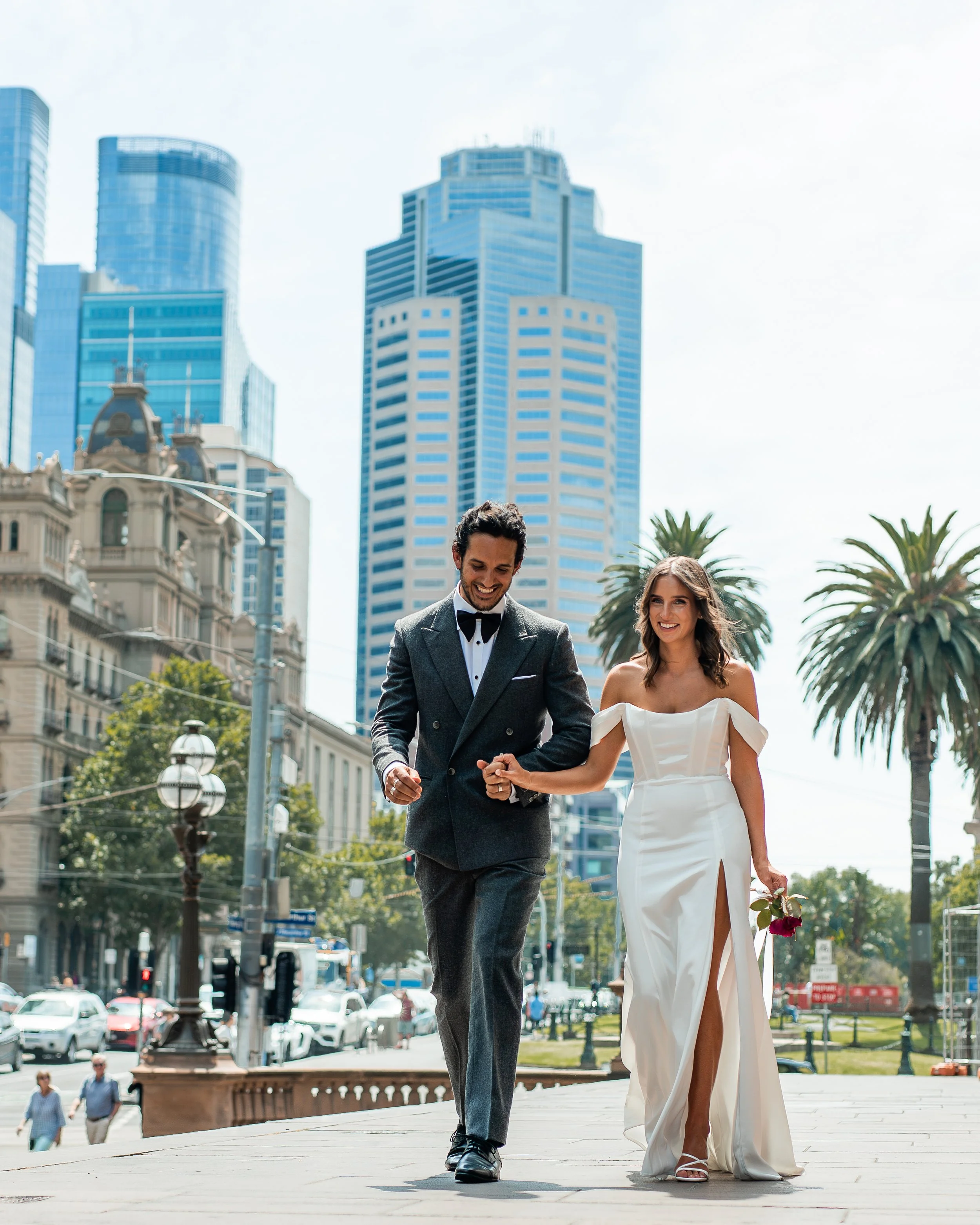 A happy couple in formal wedding attire walking outdoors in an urban setting with buildings, trees, and city life in the background.