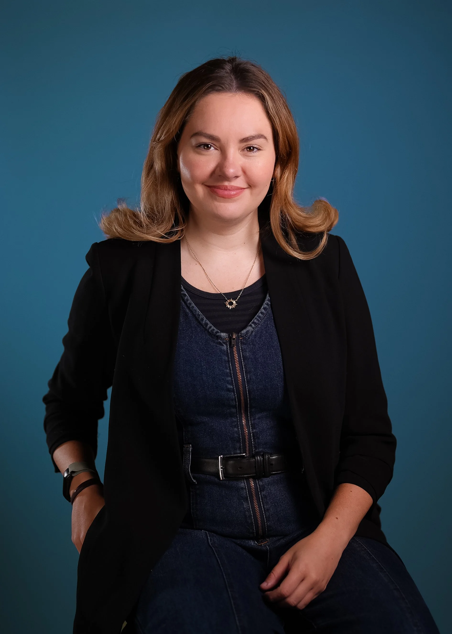 A young woman with shoulder-length light brown hair and a friendly expression sitting against a solid blue background. She is wearing a black blazer over a dark blue top, accessorized with a star-shaped necklace. One arm is resting on her knee, and the other is at her side.