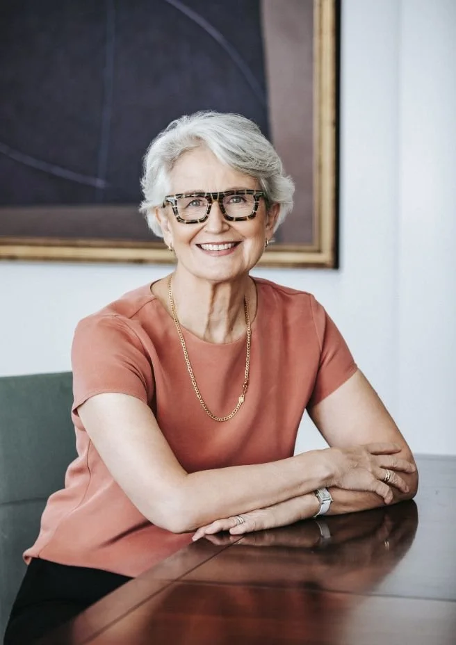 Catherine Andersen - with short gray hair and glasses sitting at a wooden table, smiling at the camera, wearing a terracotta-colored blouse and a gold necklace.