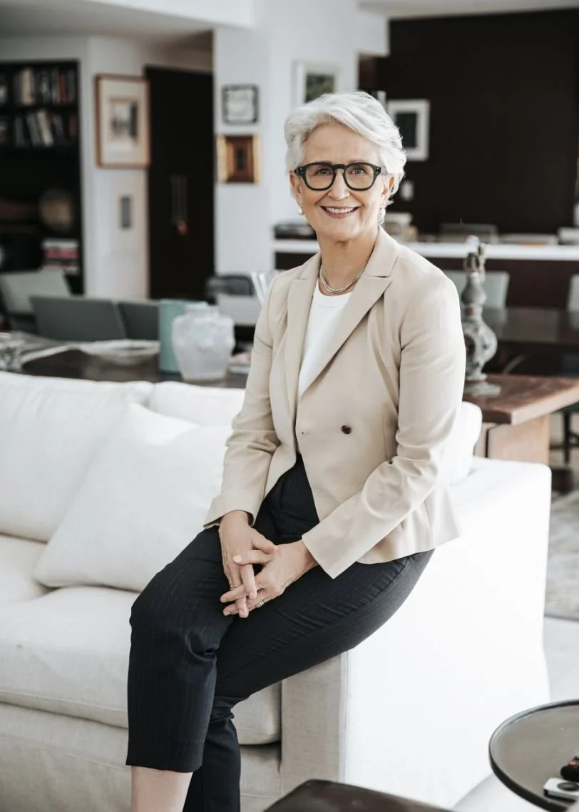 Catherine Andersen - with short white hair, wearing glasses, a beige blazer over a white top, and black pants, sitting on a white sofa in a modern living room, smiling at the camera.