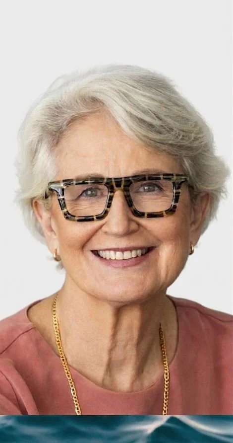 Catherine Andersen -  with short, white hair wearing patterned glasses, gold earrings, a pink top, and a gold chain, posing in front of a plain background.