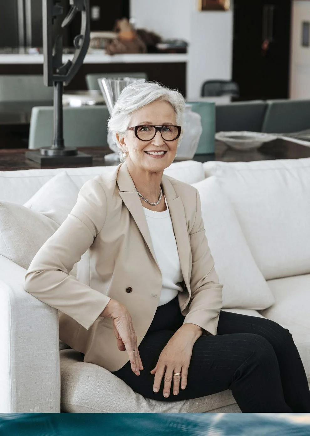 Catherine Andersen -  with short white hair, glasses, and a cheerful smile, sitting on a white sofa in a modern living room. She is wearing a beige blazer, white shirt, and black pants.