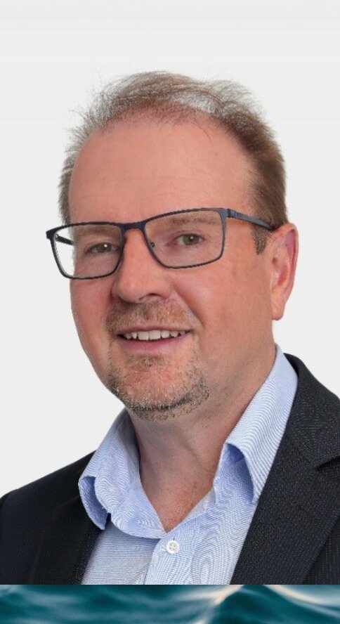 Peter Cumming - wearing glasses, with light hair, and a goatee, smiling, wearing a light blue shirt and blazer, against a white background.