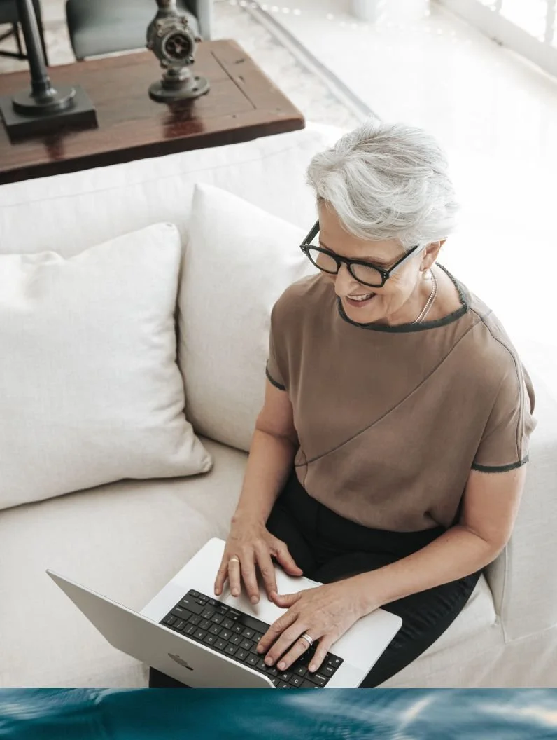 Catherine Andersen - with short gray hair and glasses smiling while sitting on a cream-colored sofa, using a silver laptop.