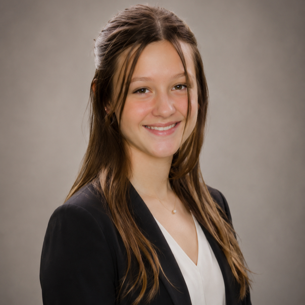 Smiling young woman with long brown hair wearing a black blazer and white top, portrait against gray background.