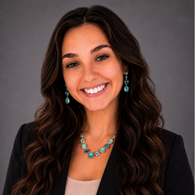 Portrait of a young woman with long, wavy brown hair, smiling, wearing a black blazer and turquoise jewelry, standing against a dark gray background.