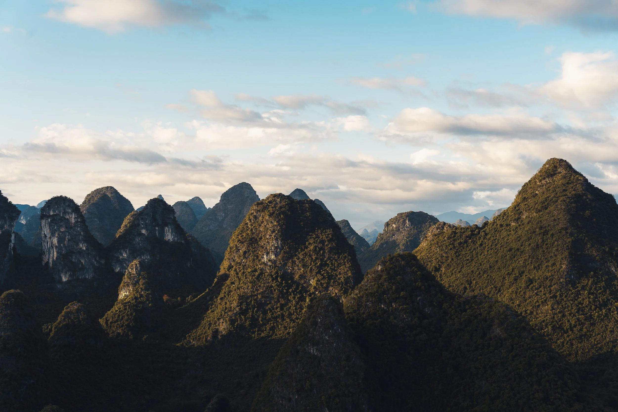 A landscape view of multiple lush green mountain peaks under a partly cloudy sky.